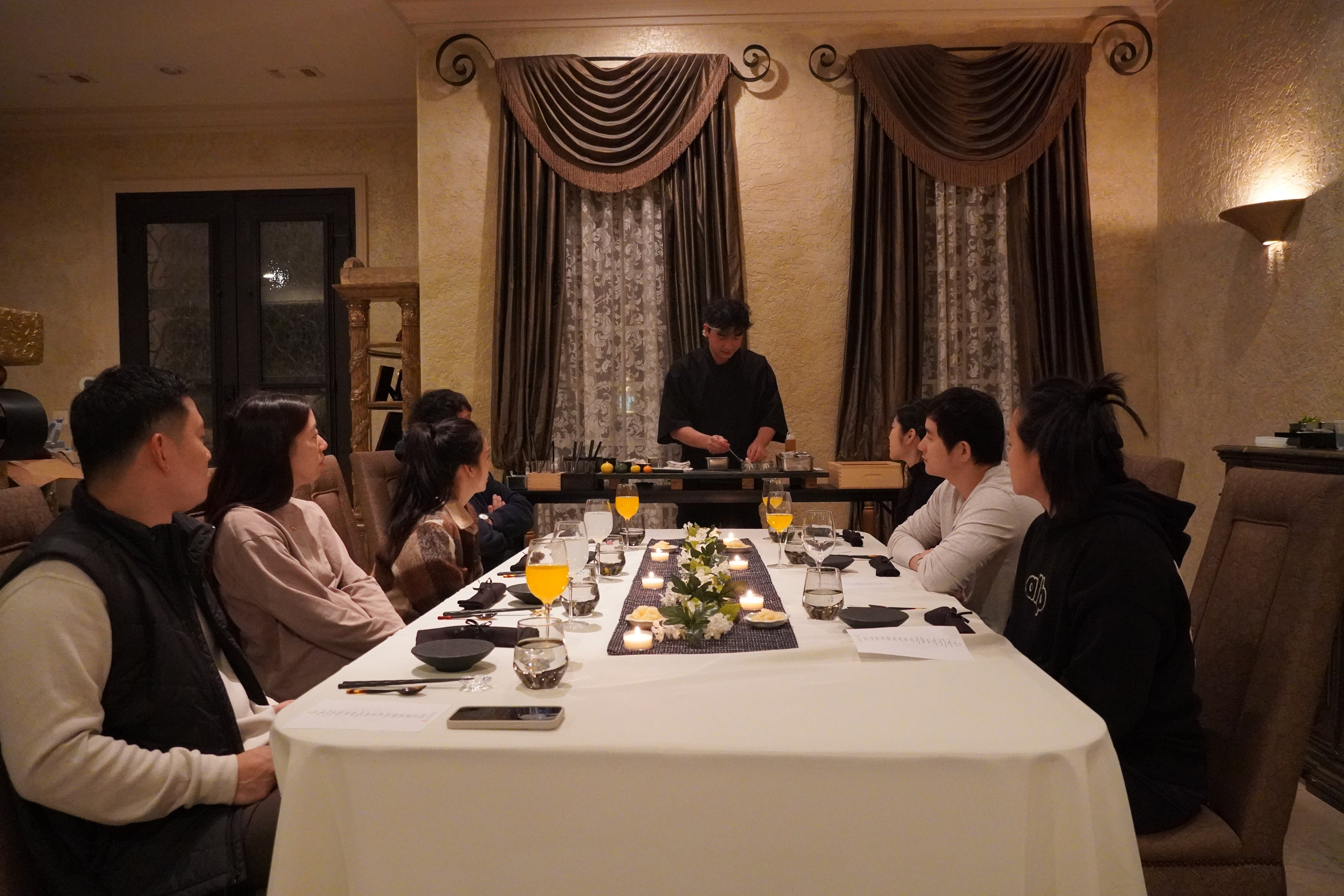 Group of people sitting around a dining table in a dimly lit room with Chef Yayo giving them the best omakase experience in their life.