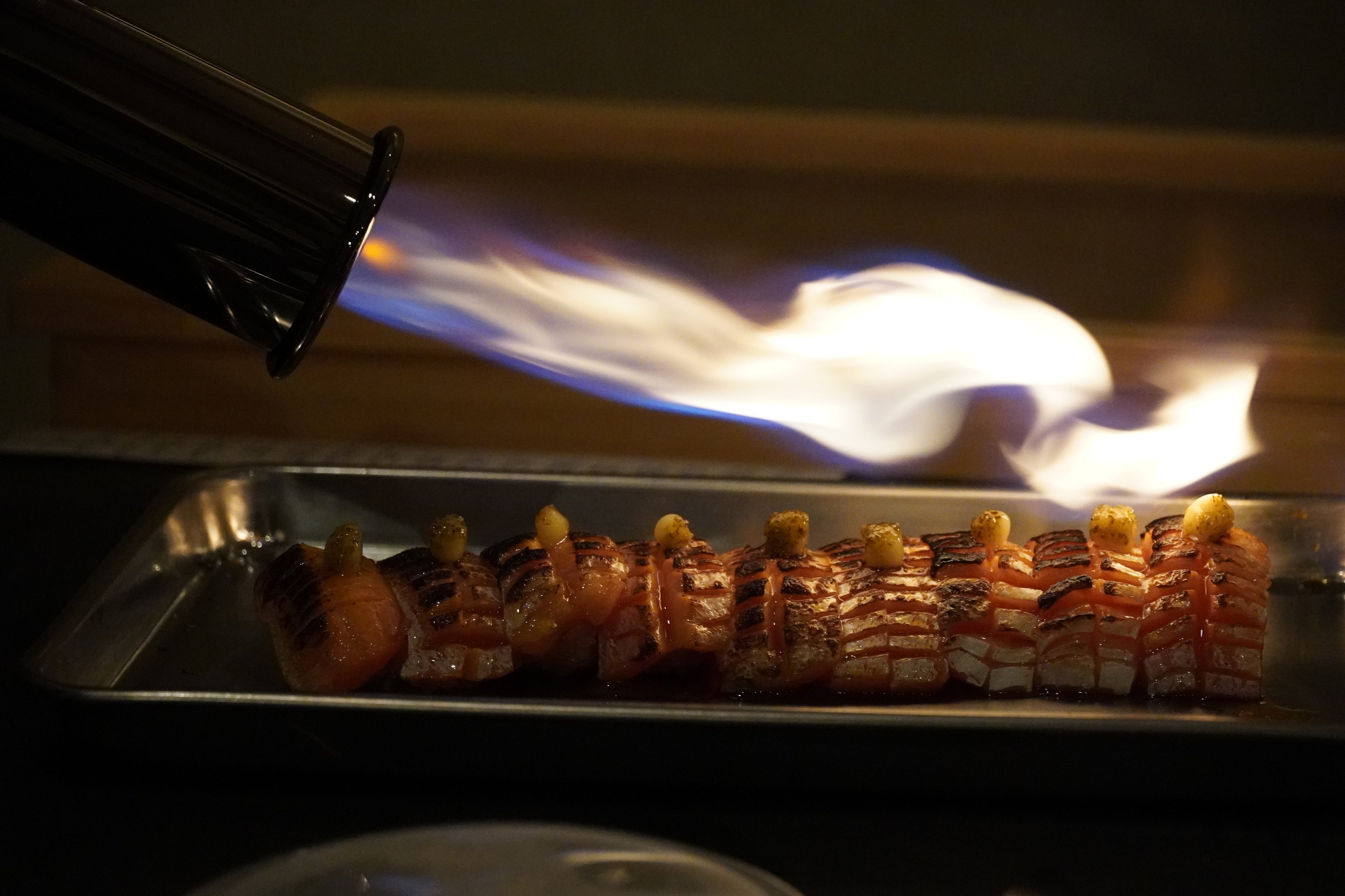 Flaming torch being used to cook sushi on a metal tray.